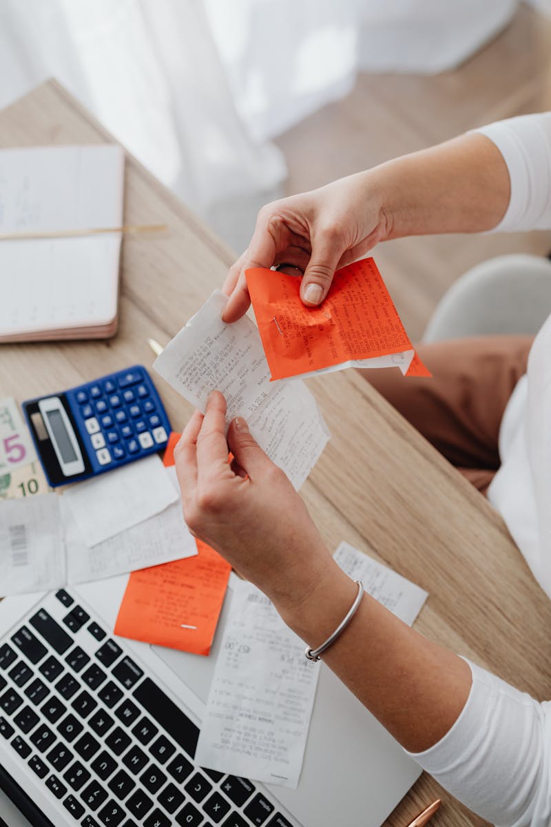 Hands sorting through receipts and notes at a desk with a calculator and laptop